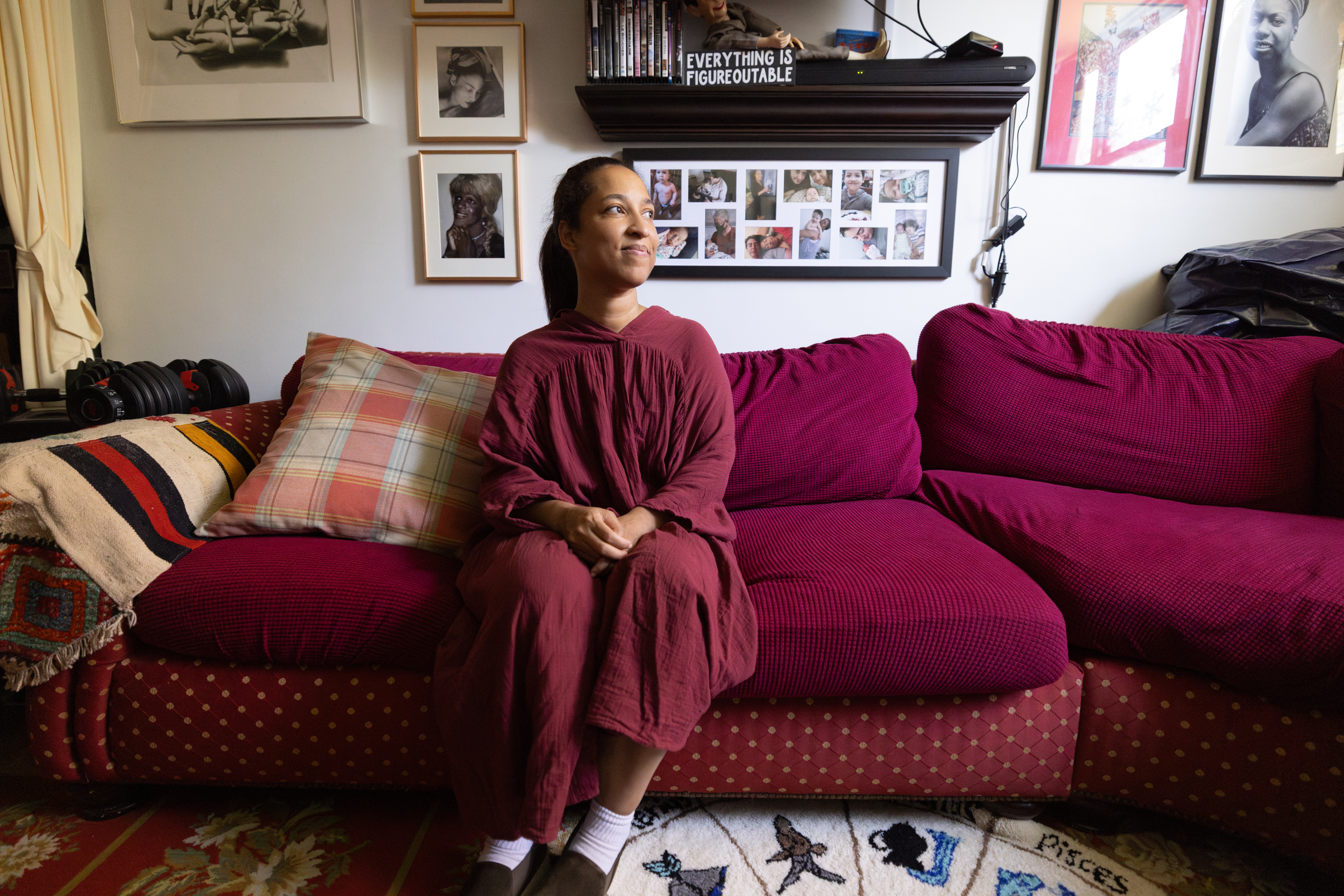 Woman smiles and sits on couch looking toward window. Framed photos are behind her. 